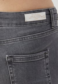 Gros plan sur un jean en denim gris, avec une poche arrière et une étiquette blanche avec du texte, cousue avec un fil de couleur claire. Texture lisse et robuste.