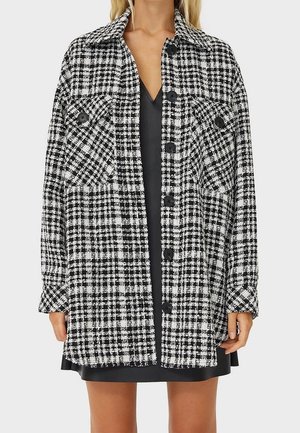 Black and white checkered long-sleeve button-up jacket with two front flap pockets worn over a black dress.
