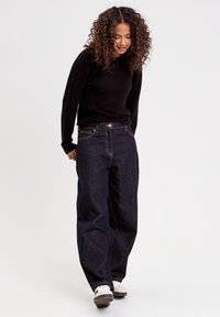 Jean large en denim foncé avec taille haute, présentant un style classique à cinq poches et une couture de contraste subtile. Porté avec un pull noir texturé.