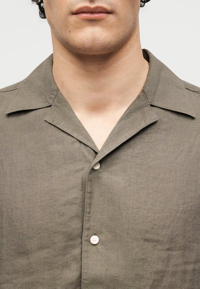 Άνδρας που φοράει ένα πουκάμισο χακί με κουμπιά και γιακά τύπου κάμπινγκ, εμφανίζεται από τον λαιμό έως το μέσο του στήθους σε απλό φόντο.