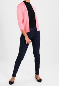 Jasnoróżowy kardigan z rękawami do trzech czwartych noszony na czarnym golfie, zestawiony z ciemnoniebieskimi obcisłymi dżinsami i czarnymi balerinami.