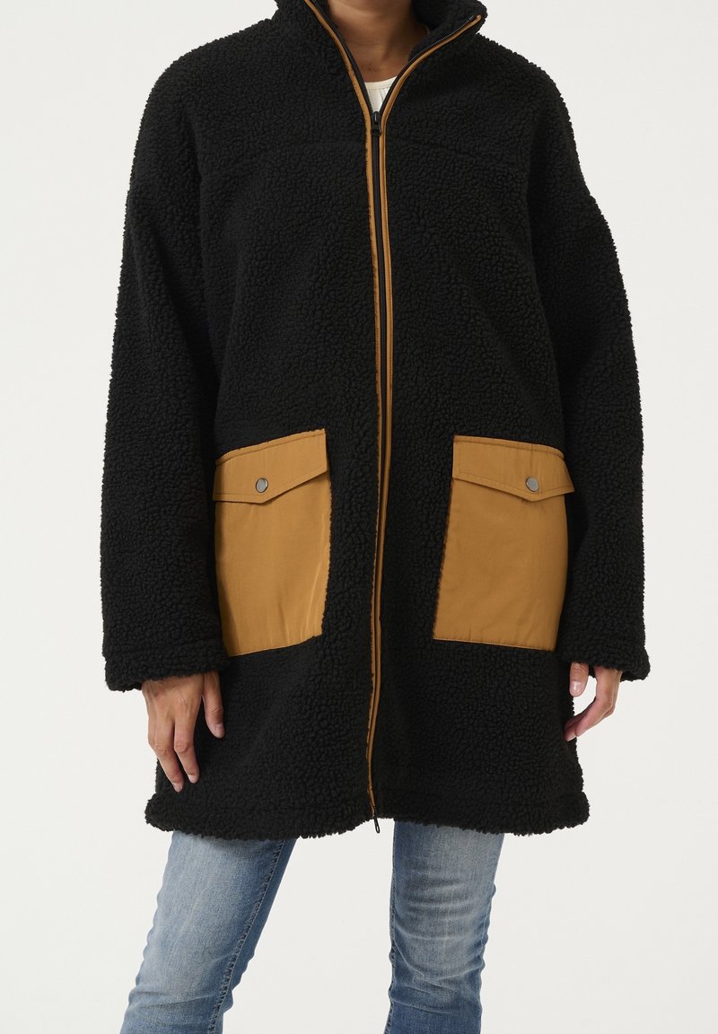 Czarny płaszcz z polaru z zamkiem z przodu, z dwoma beżowymi kieszeniami na guziki i wysokim kołnierzem. Tkanina ma teksturowany, puszysty wygląd.