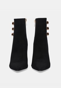 Botas de tobillo de ante negro con puntera puntiaguda, que cuentan con una cremallera lateral y tres acentos circulares dorados en el lado exterior.