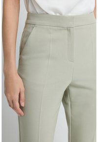 Close-up van een persoon die lichtbeige op maat gemaakte broeken met zijzakken en een witte ingestopte top draagt.