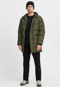 Tamnozeleni pufferjakna s kapuljačom, s prednjim zatvaračem, dva bočna džepa i pikasta tekstura, nosi se preko crne majice i hlača.