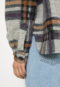 Fleece overhemd met knopen heeft een grijze basis met zwarte, oranje en paarse ruitpatronen. De manchetten zijn voorzien van een zwarte knoopdetail.