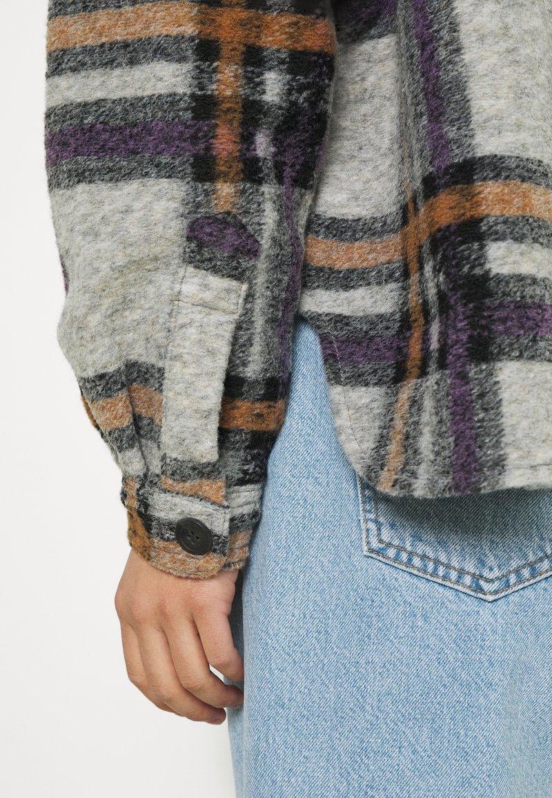 Fleece overhemd met knopen heeft een grijze basis met zwarte, oranje en paarse ruitpatronen. De manchetten zijn voorzien van een zwarte knoopdetail.