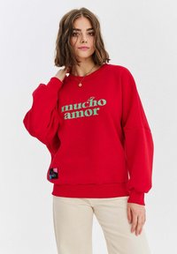 Czerwona bluzka z grafika "mucho amor" w odcieniu teal. Posiada luźny krój, ściągacze przy mankietach i dolnej krawędzi. Metka widoczna z dolnej strony. Gładkie tło.