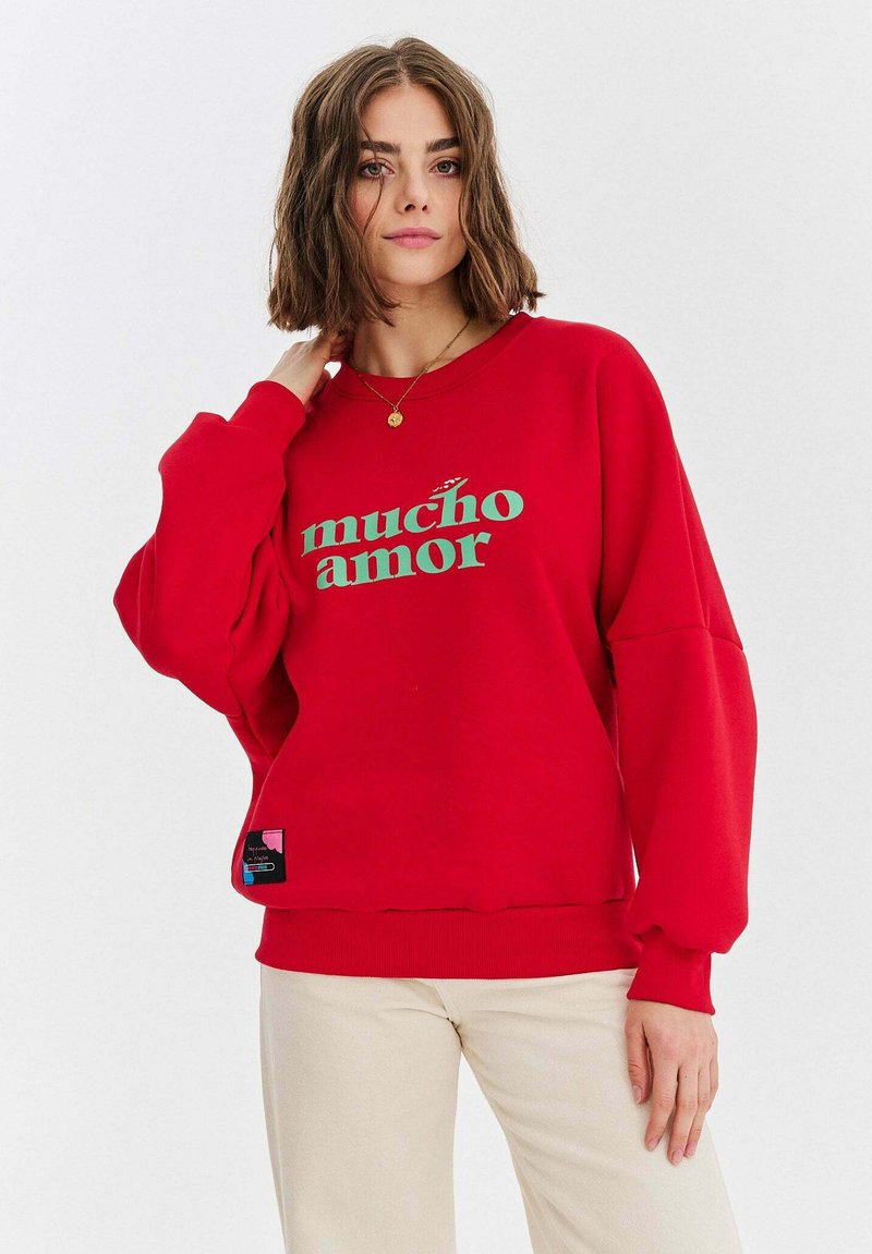 Czerwona bluzka z grafika "mucho amor" w odcieniu teal. Posiada luźny krój, ściągacze przy mankietach i dolnej krawędzi. Metka widoczna z dolnej strony. Gładkie tło.