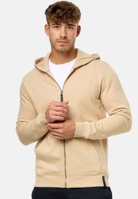 Beige zip-up hoodie i blødt materiale med hætte, frontlommer, ribbede manchetter og et lille logo på brystet.