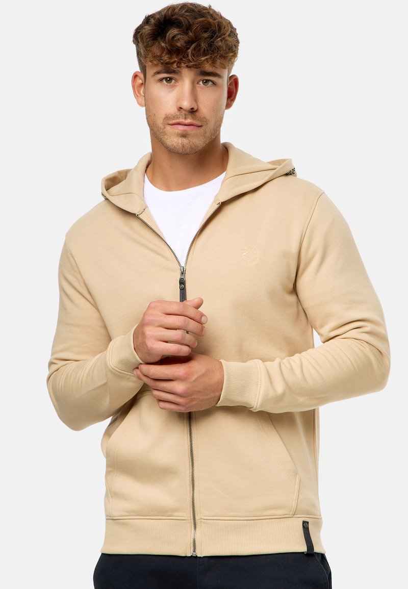 Beige Zip-Hoodie aus weichem Material, mit Kapuze, frontalen Taschen, gerippten Bündchen und einem kleinen Logo auf der Brust.