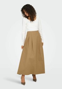 Jupe longue taille haute marron avec des plis, réalisée en tissu léger. Associée à un top blanc ajusté à manches longues et des talons gris.