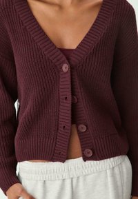 Bordeauxfarbene Strickjacke mit tiefem V-Ausschnitt und Rippendetails, ausgestattet mit fünf runden Knöpfen und einer verkürzten Passform. Strukturierter Stoff mit leichtem Stretch.