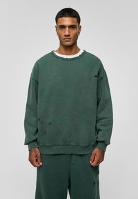 Donkergroene oversized sweatshirt van zachte stof, met versleten details, een ronde hals en geribde manchetten. Gepaard met bijpassende broeken.