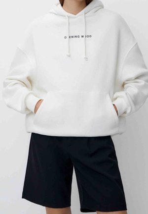 Osoba ubrana w białą oversize bluzę z kapturem z napisem „OPENING MOOD” i czarne szorty do kolan, ręce w kieszeniach bluzy.