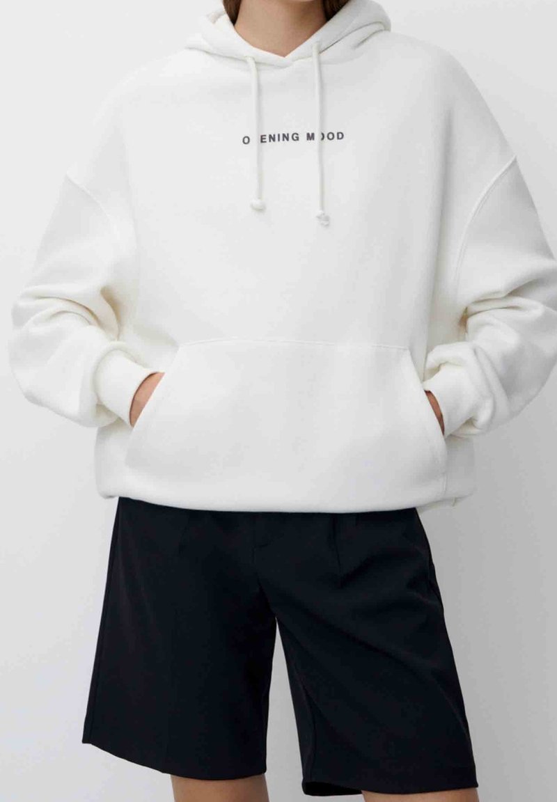 Personne portant un sweat à capuche blanc oversize avec le texte « OPENING MOOD » et un short noir jusqu’aux genoux, mains dans les poches du sweat.