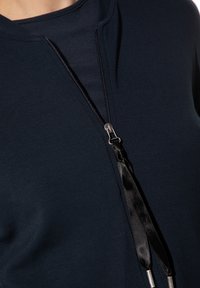 Marineblaue Bluse mit einem Reißverschluss, ausgestattet mit schwarzen satinierten Zugbändern und silbernen Hardware-Details für zusätzlichen Stil und Funktionalität.