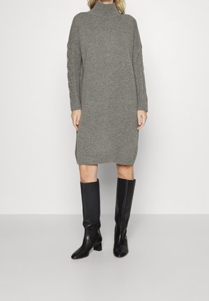 Szara dzianinowa sukienka swetrowa z golfem o długości do kolan z rękawami w warkocze, noszona z czarnymi wysokimi kozakami na obcasie.