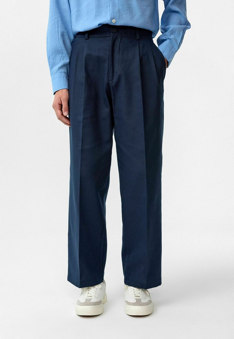 Marineblauwe, geplooide broeken met een relaxed fit. Gemaakt van een soepele stof, met riemlussen en zijzakken. Gepaard met witte sneakers.