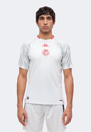Jonge man met een wit sportshirt en shorts met rode accenten en het embleem van het Tunesische voetbalteam, vooruit kijkend voor een egale achtergrond.