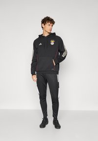 Fekete sportkapucnis pulóver, elülső zsebbel, fehér Adidas csíkokkal az ujjakon, és csapatlogóval. Párba állítva fekete, passzoló tréningnadrággal, fehér részletekkel.