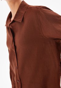 Brown linen shirt with a collar, short sleeves, and button details on the front. The fabric appears textured and lightweight.