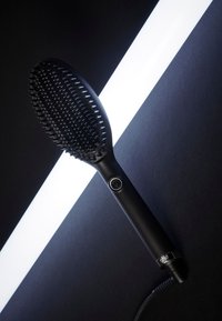 Zwarte elektrische haar stijlborstel met aan/uit-knop en kabel, diagonaal geplaatst op een donkere ondergrond met een heldere witte diagonale verlichting.