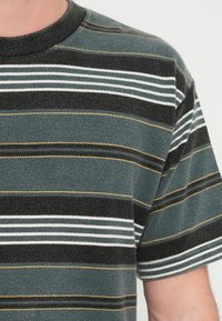 Ριγέ μπλουζάκι με οριζόντιες γραμμές σε σκούρο πράσινο, μαύρο, άσπρο και κίτρινο. Μαλακή υφή με στρογγυλή λαιμόκοψη και κοντά μανίκια.