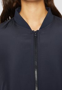 Námořnická bomber bunda má zipové zapínání, žebrovaný límec a hladkou texturu látky. Design zahrnuje kontrastní šití podél zipu.