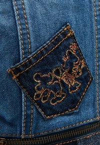 Džínová kapsa se vzorem vyšitým červenou a zelenou nití. Tmavě modrá látka, viditelné švy a na spodním okraji zip.