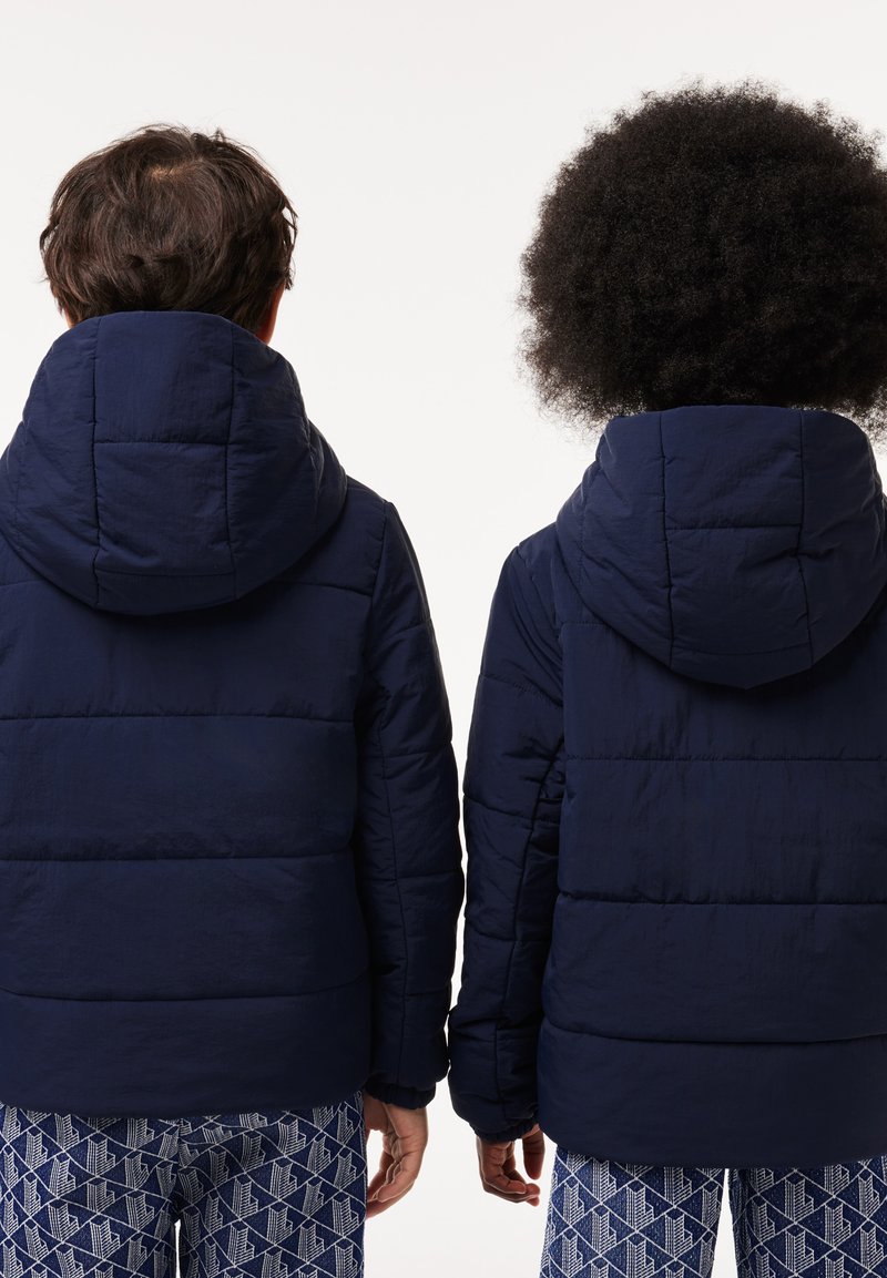 Marineblaue Steppjacken mit Kapuzen, die horizontale Nähte und eine glatte Textur aufweisen, getragen von zwei Personen, die von hinten sichtbar sind.