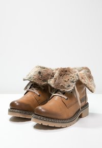 Bottes en cuir marron montant à la cheville avec doublure en fourrure repliée sur le dessus et lacets beige sur une surface blanche.