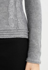 Suéter gris de punto con un patrón de trenzas a lo largo del costado, con un acabado texturizado y mangas ajustadas, combinado con una prenda negra.