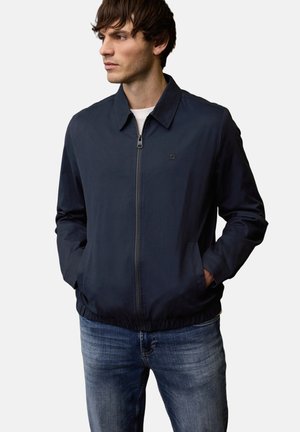 Mand iført navy lynlåsjakke med krave og blå jeans, hænderne i lommerne, kigger til siden mod en ensfarvet hvid baggrund.