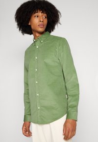 Cămașă verde din catifea cu guler button-down, având butoane frontale, mâneci lungi și o tivă curbă. Țesătură texturată.