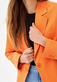 Blazer arancione realizzato in tessuto leggero, con revers a punta, una tasca e due bottoni. Indossato sopra un top nero con jeans blu.