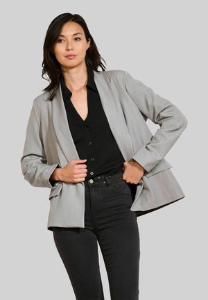 Vrouw met donker haar, gekleed in een grijze blazer, zwart overhemd met knopen en zwarte jeans, terwijl ze haar handen aan de blazer aanpast tegen een effen achtergrond.