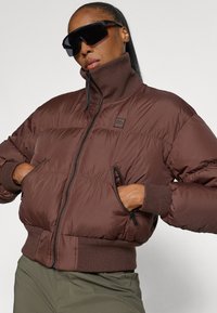 Casaco puffer castanho com colarinho alto, fecho éclair frontal e punhos canelados. Possui dois bolsos frontais com fecho éclair e um emblema.