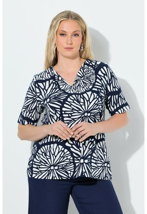 Donna bionda con capelli sciolti, indossa una camicetta con scollo a V e motivo floreale bianco e blu navy, abbinata a pantaloni blu navy, in piedi su uno sfondo bianco uniforme.