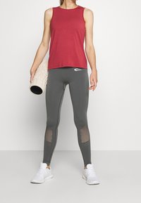 Czerwona bezrękawnikowa koszulka w połączeniu z szarymi legginsami z siateczkowymi wstawkami, uzupełniona białymi butami sportowymi oraz fakturowanym wałkiem do ćwiczeń.