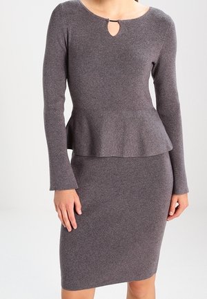 Femme portant une robe en maille grise, cintrée, à manches longues, avec un peplum à la taille et un petit détail en forme de trou à l'encolure.