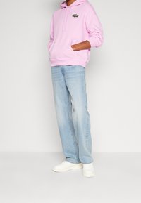 Personne portant un sweat à capuche rose clair avec un logo vert, un jean bleu clair ample et des baskets blanches, debout avec les mains dans la poche du sweat.