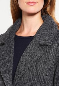 Szary teksturowany płaszcz z wyciętym kołnierzem, wykonany z grubego dzianinowego materiału. Noszony na ciemnoniebieskiej koszulce, podkreślający warstwowy look.