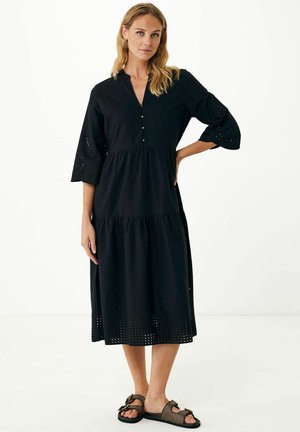 Zwarte katoenen jurk met een gelaagde ontwerp, v-hals, knoopdetails en oogjesaccenten op de mouwen en onderzoom. Gecombineerd met platte sandalen.