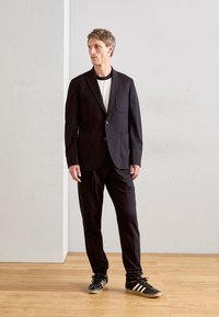 Mand i sort blazer, matchende bukser, hvid skjorte og sorte Adidas sneakers står på trægulv mod en helt hvid væg.