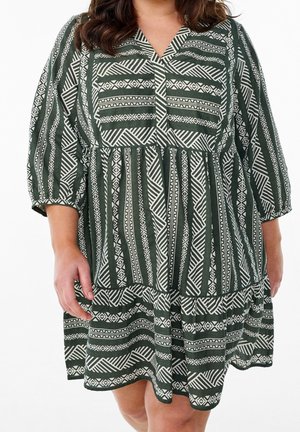 Femme portant une robe ample jusqu'aux genoux avec des motifs tribaux géométriques verts et blancs et des manches trois-quarts.