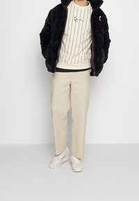 Chaqueta de piel sintética negra, sudadera de rayas crema, pantalones beige y zapatillas ligeras. Chaqueta con cremallera y capucha; la sudadera tiene un detalle bordado.