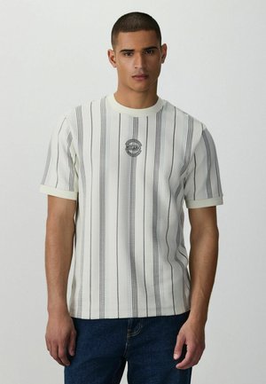 Young man wearing a white short-sleeve shirt with vertical black stripes and "Milano" logo, paired with dark blue jeans, standing against plain background.