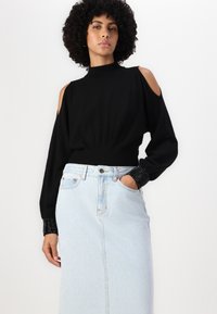 Črna pulover s prostimi rameni, z rebrastim izrezom in manšetami, v kombinaciji z svetlo modro denim krilom. Pulover ima teksturirane črne manšete.