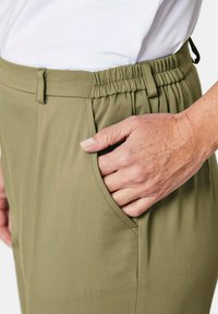 Olivgrüne Hose mit einem elastischen Bund, ausgestattet mit Seitentaschen und einer glatten Textur. Das Material wirkt leicht und flexibel.